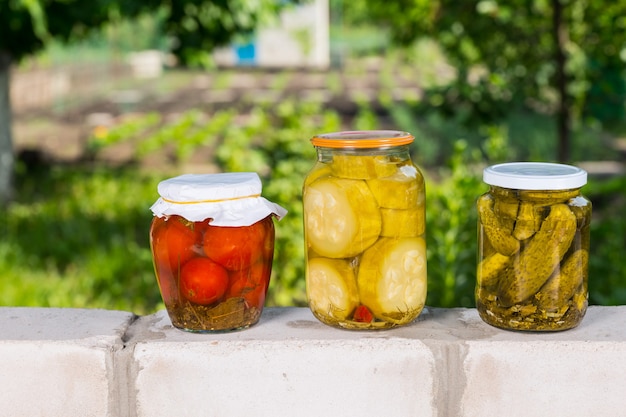 Tre vasetti di verdure appena conservate sulla sommità di un muro di mattoni - Vaso di pomodori sottaceto, cipolle e cetrioli nella parte anteriore del giardino