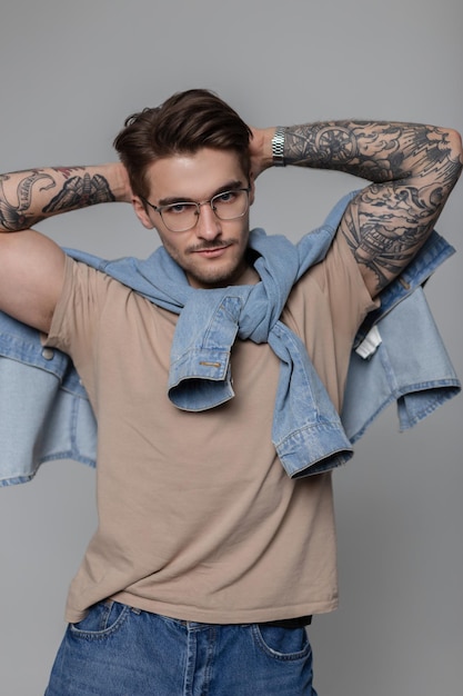 Trendy sexy hipster modello uomo con occhiali vintage che indossa abiti eleganti con una camicia di jeans e jeans e maglietta pone con le mani dietro la testa in studio e guarda la telecamera