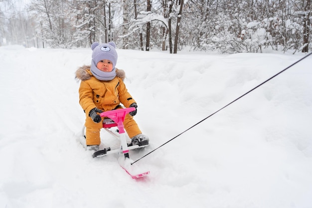 Triste bambino cavalca attraverso la neve in un parco invernale su un gatto delle nevi per bambini che si aggrappa al volante rosa