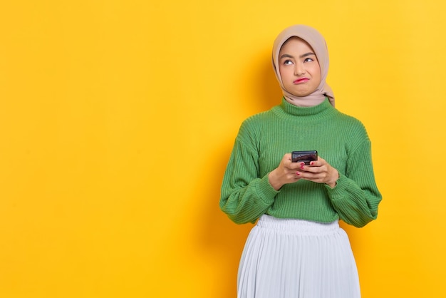 Triste bella donna asiatica in maglione verde utilizzando il telefono cellulare e guardando da parte lo spazio della copia isolato su sfondo giallo