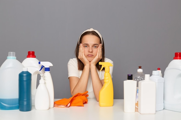 Triste cameriera adulta giovane annoiata tiene le mani sulle guance essendo stanca delle faccende domestiche indossando una maglietta bianca circondata da detergenti per la pulizia ha un'espressione triste isolata su sfondo grigio