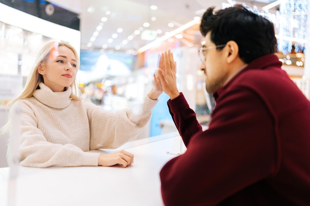 Triste coppia romantica di elegante uomo barbuto con gli occhiali e affascinante donna bionda che chiacchiera attraverso il divisorio di vetro in piedi nella sala del centro commerciale