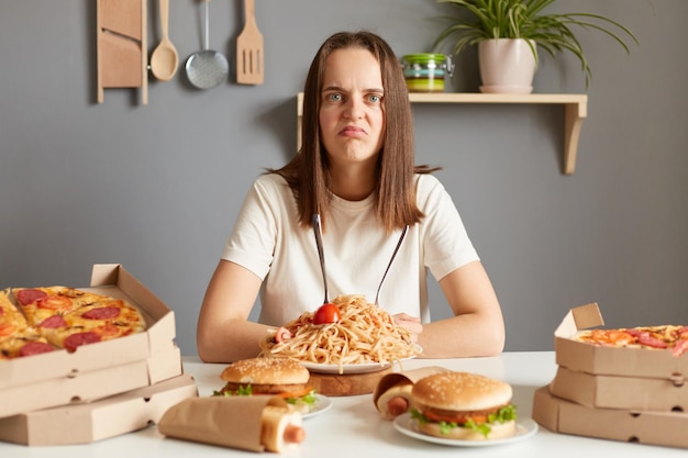 Triste donna infelice che indossa una maglietta bianca che si trova a tavola con fast food in cucina guardando la fotocamera con il viso dispiaciuto che abbatte l'eccesso di cibo nutrizione malsana