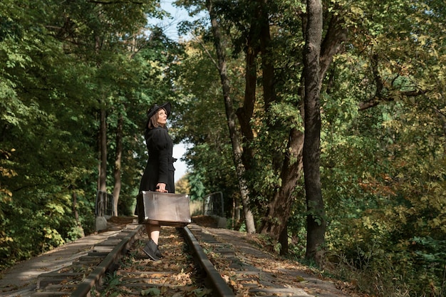 Triste donna lascia con una valigia lungo le rotaie attraverso la foresta autunnale Donna in abiti neri Vista posteriore