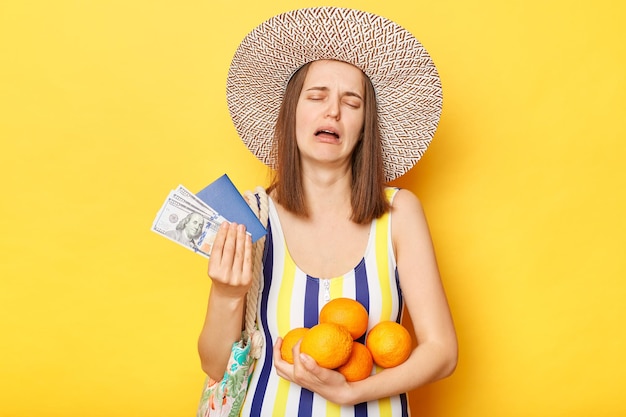 Triste donna piangente che indossa un costume intero a righe e cappello di paglia in posa isolato su sfondo giallo con in mano passaporto e documenti di frutta sconvolti