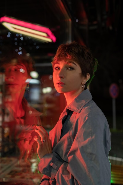 Triste donna solitaria alla luce al neon rossa appoggiata alla vetrina del negozio sulla strada di notte guardando la macchina fotografica