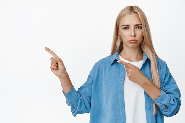 Triste e cupa donna bionda che punta il dito a sinistra nello spazio della copia accigliata sconvolto mostrando cattive notizie in piedi in abito casual su sfondo bianco