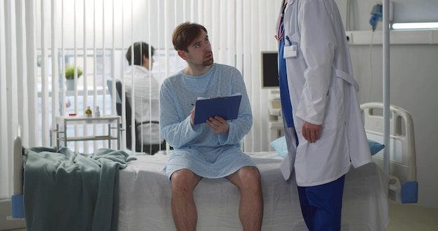 Triste giovane paziente maschio firma modulo medico seduto sul letto d'ospedale in clinica moderna