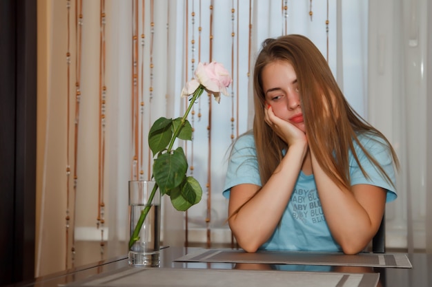 Triste piuttosto giovane donna adolescente seduta a tavola a casa con una rosa solitaria, che simboleggia la tristezza. La femmina carina mostra le emozioni della recitazione, alla finestra in cucina a casa. Spazio del copyright