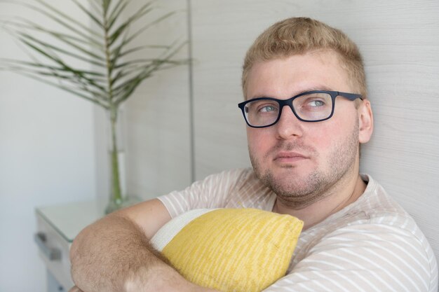 Triste premuroso giovane biondo con gli occhiali sdraiato sul divano di casa che distoglie lo sguardo abbracciando il cuscinone