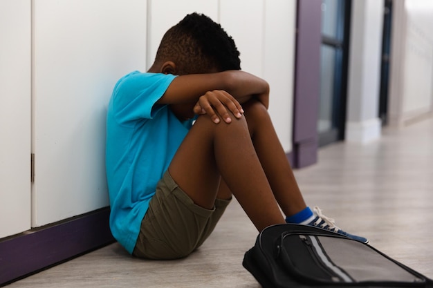 Triste scolaretto afroamericano che abbraccia le ginocchia mentre è seduto sul pavimento nel corridoio