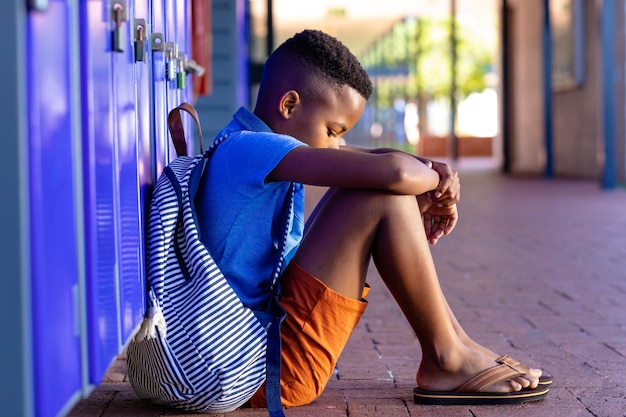 Triste scolaretto afroamericano con borsa scolastica seduto accanto agli armadietti a scuola con spazio per copie