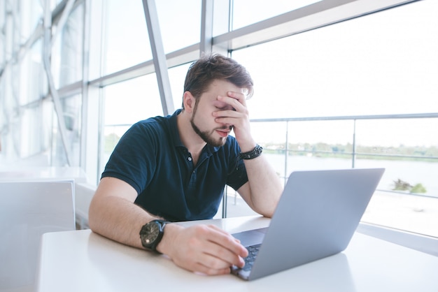 Triste uomo in abiti casual è seduto al tavolo e si chiude il viso con la mano.