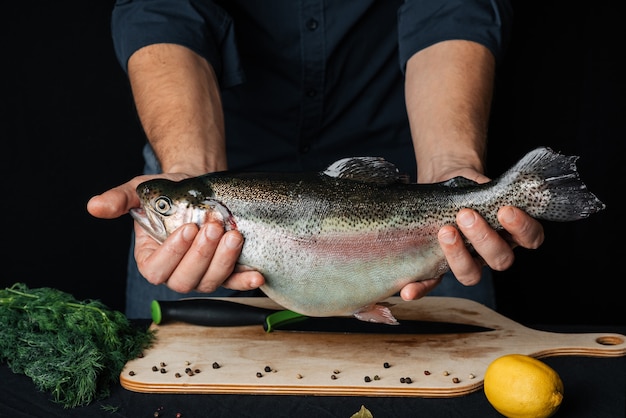 Trota iridea nelle mani di un cuoco maschio sullo sfondo della cucina. Pesce crudo fresco