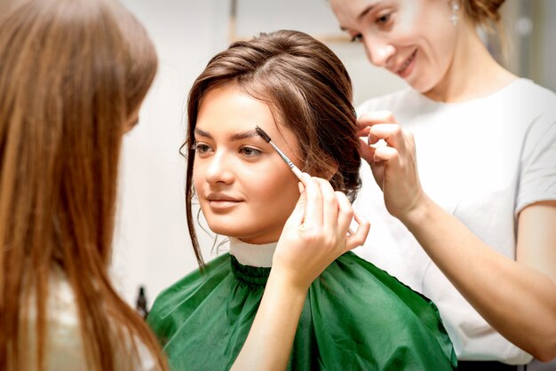 Truccatore che pettina le sopracciglia e parrucchiere preparando l'acconciatura per la bella giovane sposa caucasica in un tandem di salone di bellezza di due maestri di bellezza