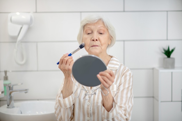 Trucco. Donna dai capelli grigi in pigiama che fa il trucco in bagno