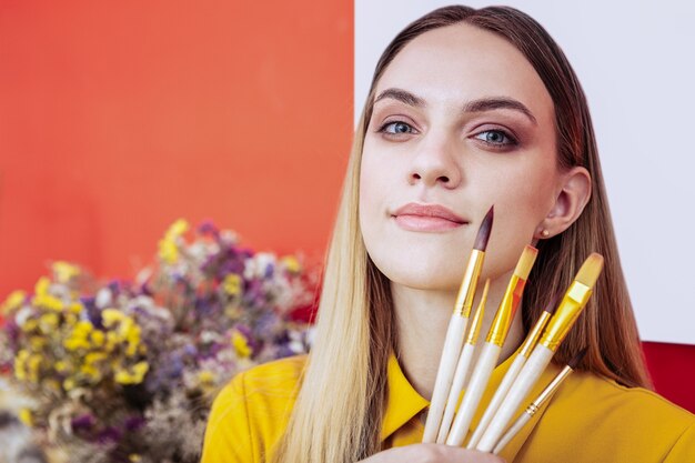 Trucco naturale. Artista bionda con trucco naturale che tiene i pennelli per pittura in piedi vicino al muro rosso