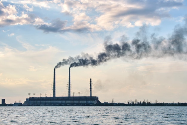 Tubi alti della centrale elettrica a carbone con fumo nero che si muove verso l'alto atmosfera inquinante sopra l'acqua del lago.