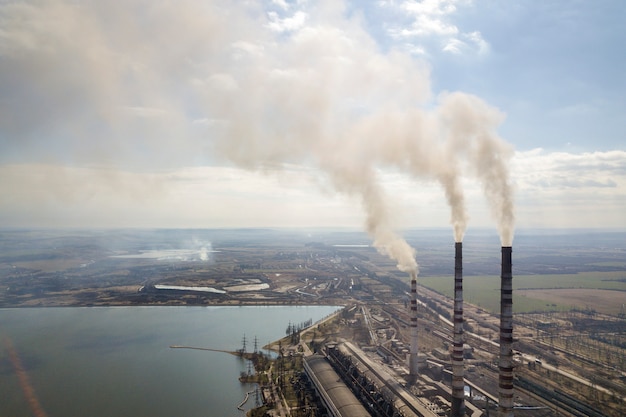 Tubi alti della centrale elettrica, fumo bianco su paesaggio rurale, acqua del lago e cielo blu.