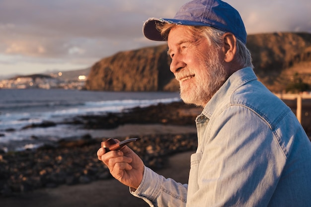 Tubo di fumo anziano bello dell'uomo barbuto che si siede all'aperto alla spiaggia al tramonto che gode rilassarsi