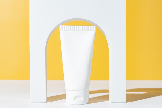 Tubo per cosmetici in plastica bianca vuota e podio ad arco bianco su sfondo giallo Trattamento di bellezza per la cura della pelle Concetto di spa Vetrina alla moda di prodotti cosmetici