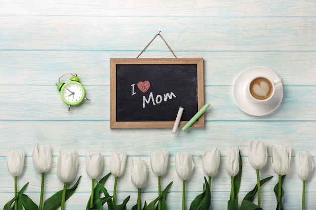 Tulipani bianchi con bordo di gesso, tazza di caffè e sveglia sui bordi di legno blu. Festa della mamma