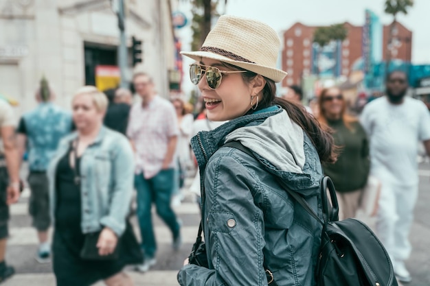 turista asiatico sorridente felice con lo zaino che cammina sull'incrocio della strada della zebra in diverse multi persone nella città urbana. vista laterale giovane ragazza con cappello di paglia e occhiali da sole rilassarsi visitando la città di hollywood