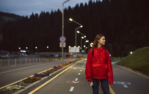 Turista attraente della ragazza in un impermeabile rosso che osserva da parte camminando sulla strada in montagna