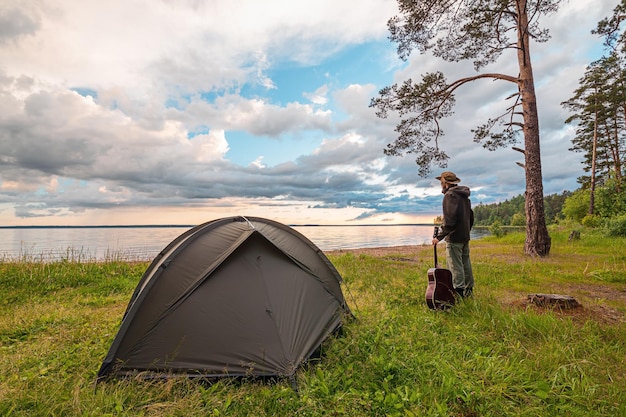 Turista con la chitarra in piedi vicino alla tenda del campo sulla riva del lago godendosi la vista al tramonto