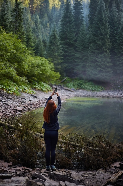 Turista della donna capelli rossi che cattura foto utilizzando il suo smartphone vicino al lago di montagna. Escursioni nelle montagne dei Carpazi, Lago Rosohan