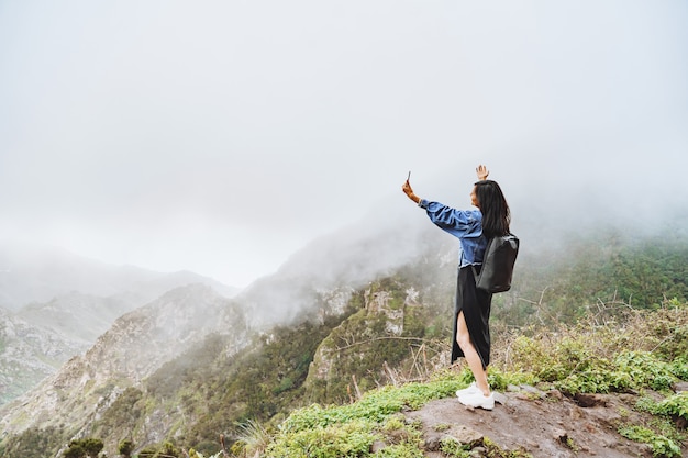 Turista della donna felice che prende selfie facendo uso dello smartphone sopra una montagna