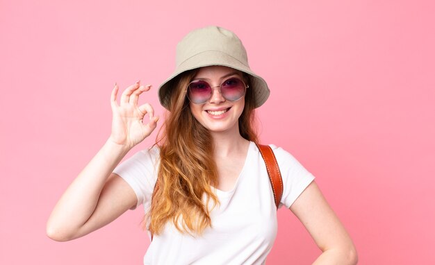 Turista della donna graziosa dalla testa rossa che si sente felice, mostrando approvazione con un gesto ok
