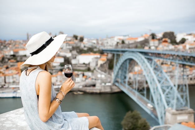 Turista della giovane donna che si siede con un bicchiere di vino di Porto sulla terrazza con una splendida vista del paesaggio urbano sulla città di Porto in Portogallo