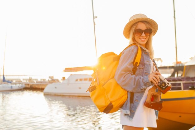 Turista della giovane donna con il grande zaino da viaggio e la macchina fotografica all'alba allo yacht Club. Concetto di viaggio e turismo