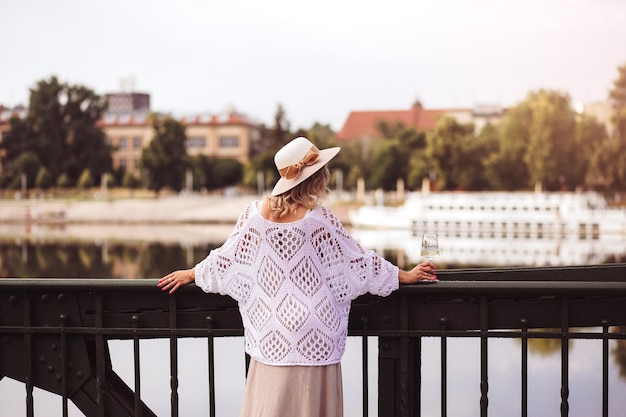 Turista della giovane donna in piedi con un bicchiere di vino bianco al ponte nel fiume della città europea sullo sfondo