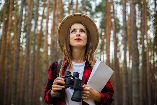 Turista della giovane donna in un cappello, camicia a quadri rossa tiene una mappa e un binocolo nella foresta.