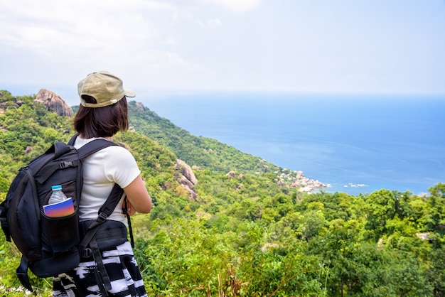 Turista delle donne con uno zaino indossare il berretto in piedi guarda al bellissimo paesaggio naturale mare blu sotto il cielo estivo dall'alto punto panoramico a Koh Tao a Surat Thani, Thailandia