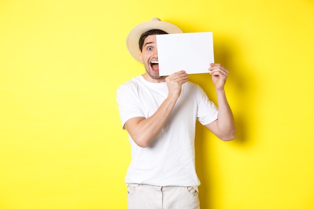 Turista eccitato in vacanza che mostra un pezzo di carta bianco per il tuo logo, con in mano un cartello vicino al viso e sorridente, in piedi su sfondo giallo