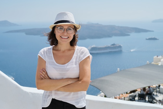 Turista felice della donna matura che viaggia sulla famosa isola di Santorini, femmina con le braccia incrociate guardando la telecamera, sfondo del mare, navi da crociera, cielo, spazio di copia