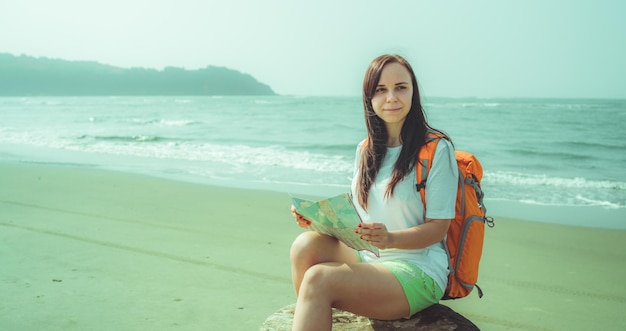 Turista femminile che legge la mappa vicino al mare Donna a piedi nudi con lo zaino seduto su legni e esamina la mappa mentre riposa sulla spiaggia sabbiosa vicino al mare ondeggiante