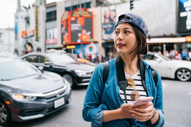 turista femminile che trova il negozio che sta facendo una grande vendita dalla sua mappa nello smartphone. Ragazza che usa l'app taxi sul cellulare. Signora asiatica in cappuccio che cammina nella via di Hollywood