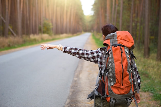 Turista femminile in una camicia a quadri con un grande zaino arancione vicino a un'autostrada nel bosco vota per ottenere un passaggio. Autostop, turismo domestico. Zaino in spalla, avventura da solo, viaggio, escursione