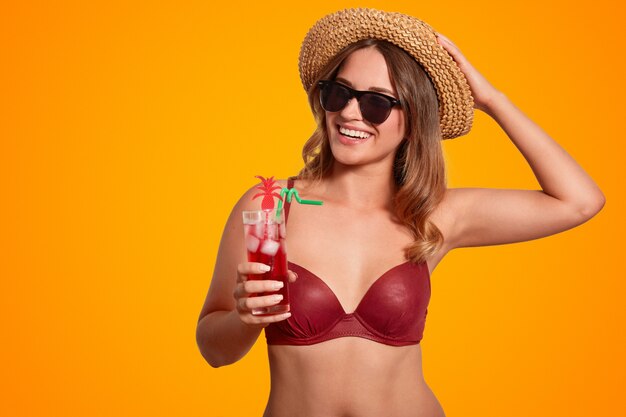 turista femminile piuttosto allegro in cappello e bikini estivo di paglia alla moda, indossa occhiali da sole alla moda, gode di un drink estivo