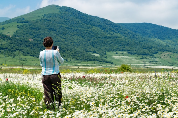 Turista femminile scattare foto con tavoletta digitale in piedi nel bellissimo campo pieno di fiori che sbocciano davanti alle montagne in Armenia