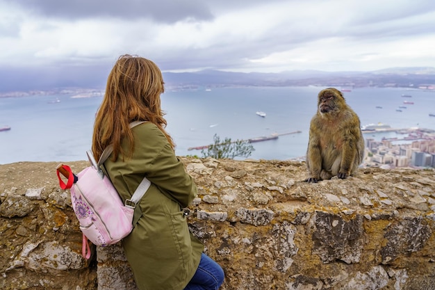 Turista femminile sulla schiena che guarda una piccola scimmia di Gibilterra in alto sulla roccia con la baia sullo sfondo