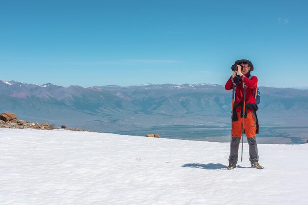 Turista in rosso cammina sulla montagna di neve vicino al bordo dell'abisso in alta quota sotto il cielo blu in una giornata di sole Uomo con la macchina fotografica sulla montagna innevata vicino al bordo del precipizio con vista sulla grande catena montuosa in lontananza