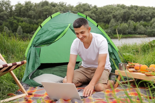 Turista maschio bruna che lavora su un computer portatile all'aperto in un campeggio giovane ragazzo freelance che lavora online su un computer portatile durante un'escursione vicino al fiume all'aperto La possibilità di lavorare da qualsiasi parte del mondo