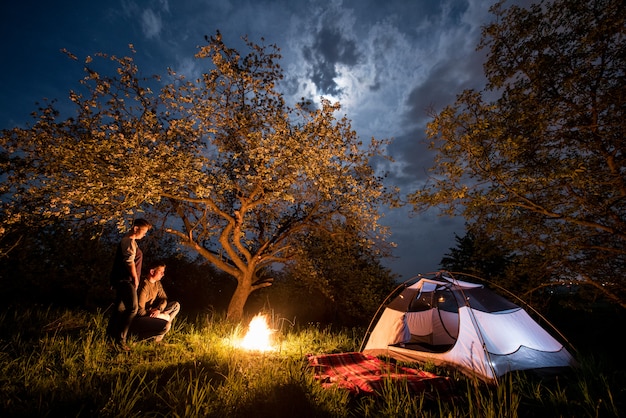 Turisti romantici delle coppie che stanno ad un fuoco di accampamento vicino alla tenda sotto gli alberi e cielo notturno con la luna. Notte in campeggio