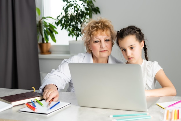 Tutore donna o madre adottiva che aiuta una ragazza caucasica carina a fare i compiti seduta al tavolo. Tata e bambino diversi che imparano a scrivere sul taccuino che studiano a casa.