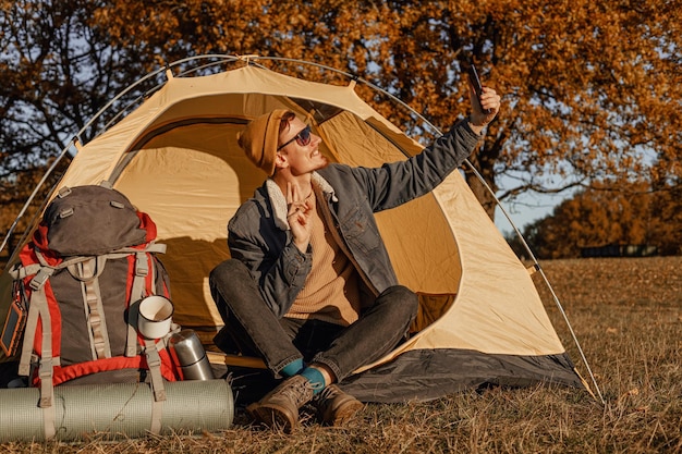 Tutta la lunghezza del viaggiatore maschio felice seduto vicino alla tenda da campeggio e allo zaino mentre si fa selfie